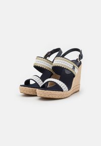 Sandalias cuña en color navy con tiras trenzadas en blanco y dorado, plataforma envuelta en yute y detalle de correa ajustable en el tobillo.