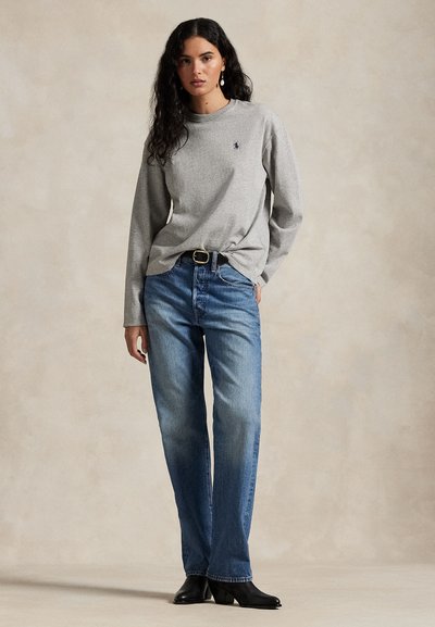 Donna con capelli lunghi e ricci che indossa una maglietta grigia a maniche lunghe, jeans blu, stivali neri e orecchini di perle, in piedi davanti a uno sfondo neutro semplice.