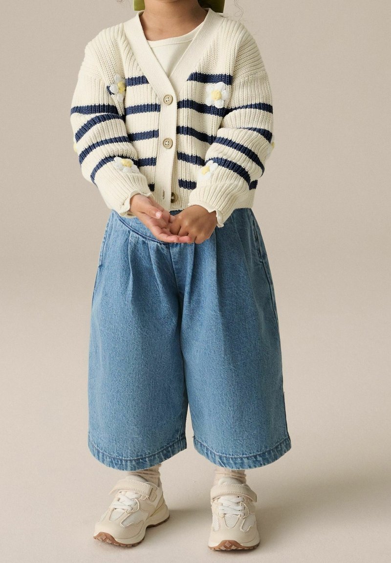 Enfant portant un cardigan crème avec des rayures et des fleurs bleu marine, un pantalon large en denim bleu, des chaussettes beiges et des baskets blanches, les mains jointes devant lui.