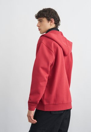 Młody mężczyzna o kręconych włosach, ubrany w czerwoną bluzę z kapturem i czarne spodnie, stojący bokiem na jednolitym jasnym tle.