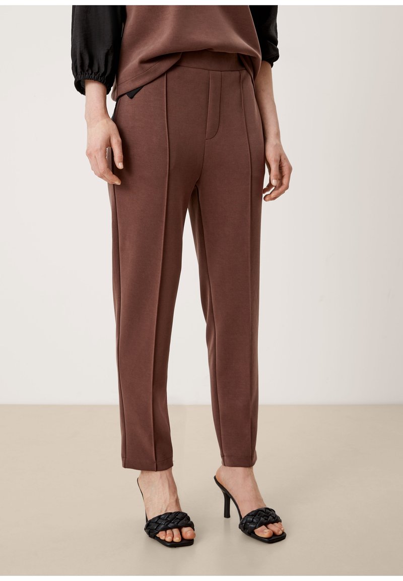 Femme portant un pantalon marron sur mesure et des sandales tressées à talons hauts noires, debout sur un sol beige devant un fond clair uni.