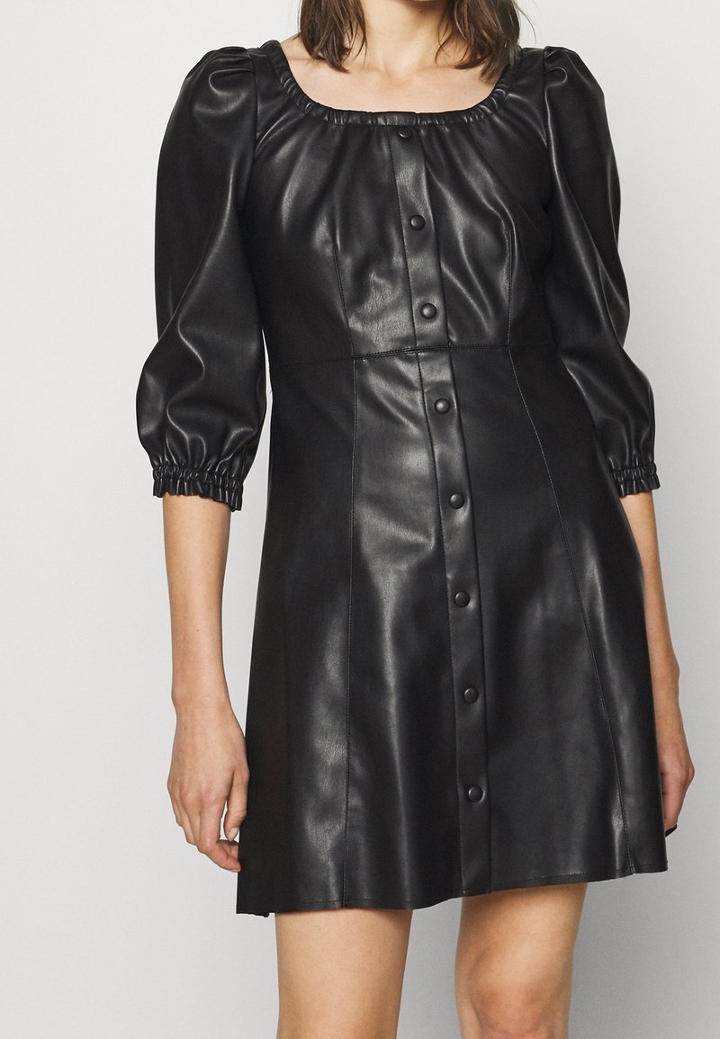 Femme portant une robe noire en similicuir avec des manches bouffantes aux coudes et des détails de boutons sur le devant, sur un fond uni.