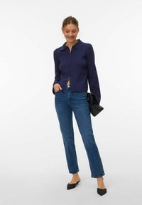 Pull en maille bleu marine à fermeture éclair avec col, associé à un jean bleu droit et des chaussures noires à bout pointu. Tenant une pochette noire.