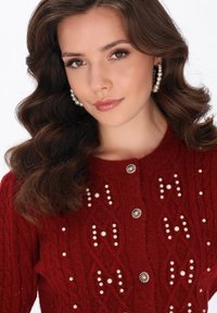 Punainen neulecardigan, joka sisältää kaapelikudontakuviota, koristeltu helmikoristeilla ja koristeellisella nappisuljennalla. Herkkää yksityiskohtaisuutta kauttaaltaan.