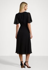 Black dress with sheer flutter sleeves and a fitted bodice. A knee-length skirt flows gently, featuring a closed back and subtle zipper.