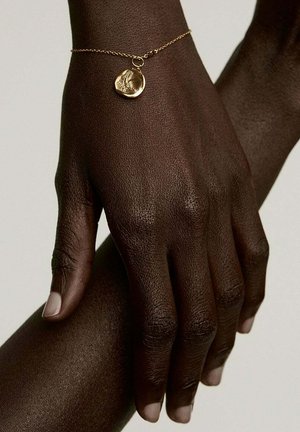 Mano de piel oscura descansando sobre el antebrazo, llevando una delicada pulsera de cadena de oro con un charm redondo grabado.