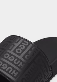 Black slide sandal with embossed repeating "HUGO" logo pattern on the strap, featuring a textured footbed and sole.