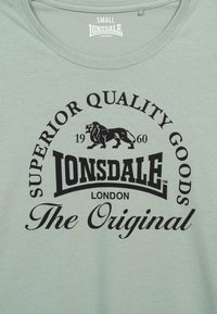 T-shirt in cotone verde chiaro con testo nero e grafica di un leone. Il design include le parole "SUPERIOR QUALITY GOODS", "LONSDALE" e "The Original."