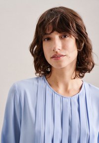 Blusa azzurro chiaro con pieghe verticali, scollo tondo e tessuto morbido e liscio; modella con capelli ricci guarda verso la fotocamera.