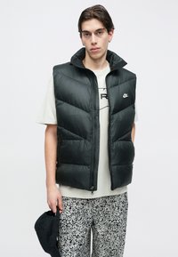 Tânăr îmbrăcat într-o vestă matlasată neagră Nike, tricou alb cu imprimeu grafic, pantaloni negri cu model alb-negru, ținând o șapcă neagră pe lângă el.