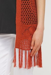Offen gestricktes orangenes Schal mit strukturiertem Muster und Fransen, getragen über einem schwarzen Oberteil mit einer Hand, die den Rand des Schals hält.