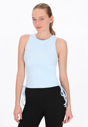 Tânără cu păr blond prins în coadă de cal, purtând un top fără mâneci, bleu deschis, cu legături laterale și pantaloni negri, stând pe un fundal alb.