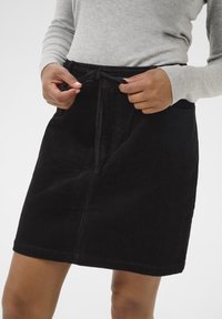 Jupe en velours côtelé noir au design droit, avec une taille à cordon de serrage et des poches latérales. La texture est côtelée et douce.