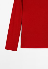 Chemise rouge à manches longues avec un ourlet droit vue de côté, mettant en avant la manche gauche et une partie du torse.