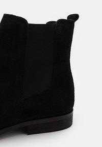 Botín negro de ante hasta el tobillo con paneles laterales elásticos, punta redondeada y un tacón de madera plano con detalles de costura sutiles.