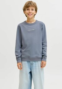 Νεαρό άτομο που χαμογελά, φοράει μπλε φούτερ με το κείμενο «Originals Studio» και ανοιχτόχρωμο τζιν με φαρδιά παντελόνα, μπροστά σε απλό φόντο.