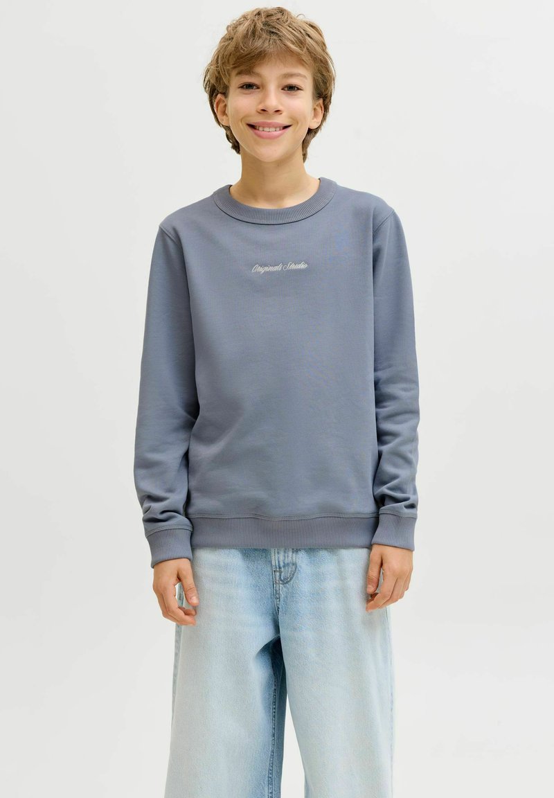 Νεαρό άτομο που χαμογελά, φοράει μπλε φούτερ με το κείμενο «Originals Studio» και ανοιχτόχρωμο τζιν με φαρδιά παντελόνα, μπροστά σε απλό φόντο.