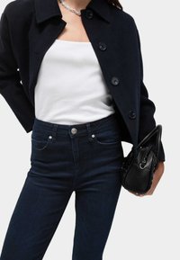 Svart ullblandad jacka med stora knappar över en vit tanktopp, i kombination med mörk denimjeans. Svart handväska med dragkedjedetalj i handen.