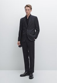 Mörkgrå skräddarsydd kostym med notchslag, enkla knappar och byxor. Svart skjorta under, håller en svart väska, står på ett ljust grått golv.