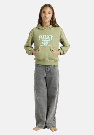 Fille debout pieds nus portant un sweat à capuche vert olive avec le logo "ROXY Endless Summer" et un jean gris ample sur un fond blanc uni.