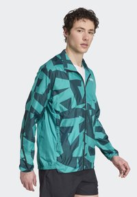 Lättvikts zip-up jacka i teal och mörkgrön med ett abstrakt geometriskt mönster. Har elastiska ärmslut och sidfickor.