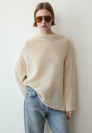 Person trägt übergroßen beigen Strickpullover, hellblaue Jeans und braune Sonnenbrille, steht vor einem schlichten hellen Hintergrund.