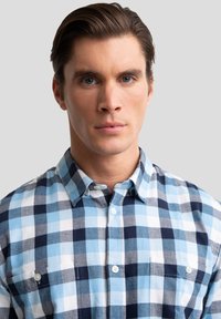 Blauw-wit geruit overhemd met knopen en kraag, met een lichte textuur en twee borstzakken, gedragen door een model tegen een neutrale achtergrond.