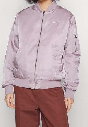 Osoba ubrana w jasną fioletową satynową kurtkę typu bomber z przednim zamkiem błyskawicznym, kieszenią na zamek na rękawie oraz białym logo Nike na piersi.