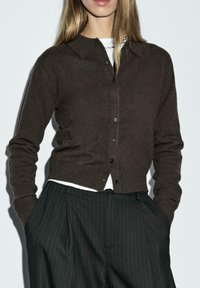 Ciemnobrązowy kardigan z miękkiego materiału, z kołnierzykiem, zapinany na guziki i z prążkowanym dołem. Połączony z spodniami w paski.