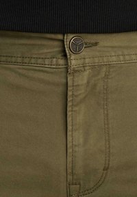 Nahaufnahme von khakifarbenen Hosen mit sichtbaren Nähten, Metallknopf mit Gravur "PME Legend" und teilweise sichtbarem schwarzem Rippenpullover am Bund.