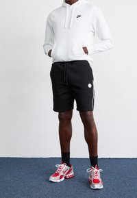 Vit hoodie med en framficka och svart Nike-logotyp, matchad med svarta shorts med vita detaljer och en cirkulär logotyp. Röda och grå sneakers.