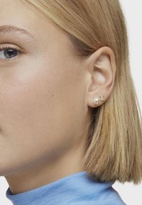 Złote kolczyki z perłą i zawieszką w kształcie misia. Projekt obejmuje gładkie i teksturowane powierzchnie, z delikatnym ogólnym wyglądem.