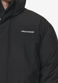 Sort isoleret jakke med høj krave, lynlåslommer og et sølvfarvet "PEGADOR" logo på brystet. Glat stof med mat overflade.