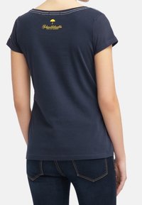 Tummansininen puuvillapaita, jossa on lyhyet hihat, pyöreä pääntie ja keltainen logo selässä. Hihansuiden kohdalla on siisti ommel.