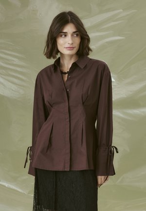 Brown button-up shirt made from cotton with a tailored fit, featuring pleats, adjustable ties at the sleeves, and a pointed collar.
