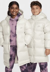 Dos personas llevan chaquetas de plumas blancas y sobredimensionadas con un diseño acolchado. Las chaquetas tienen capuchas y sutiles logotipos de Nike. Debajo, una de ellas lleva un hoodie de tie-dye púrpura y pantalones.