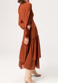 Rustinvärinen mekko pitkillä hihoilla, räystäskaitaleella ja takana solmittavalla sulkimella, yhdistettynä beigeihin polvipitkiin saappaisiin. Sileä kangas.