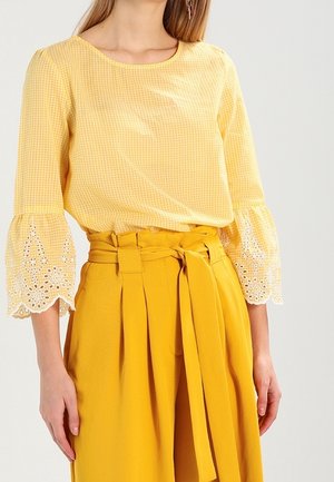 Blusa gialla a quadretti con dettagli in pizzo bianco sulle maniche a campana, abbinata a pantaloni a vita alta gialli con pieghe e cintura annodata.
