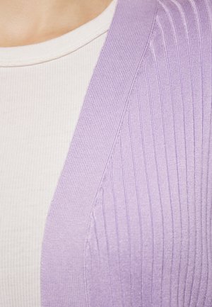 Lila geribde cardigan is over een witte, gestructureerde top met korte mouwen gedragen. De cardigan heeft een zachte, gebreide stof met een lichte glans.