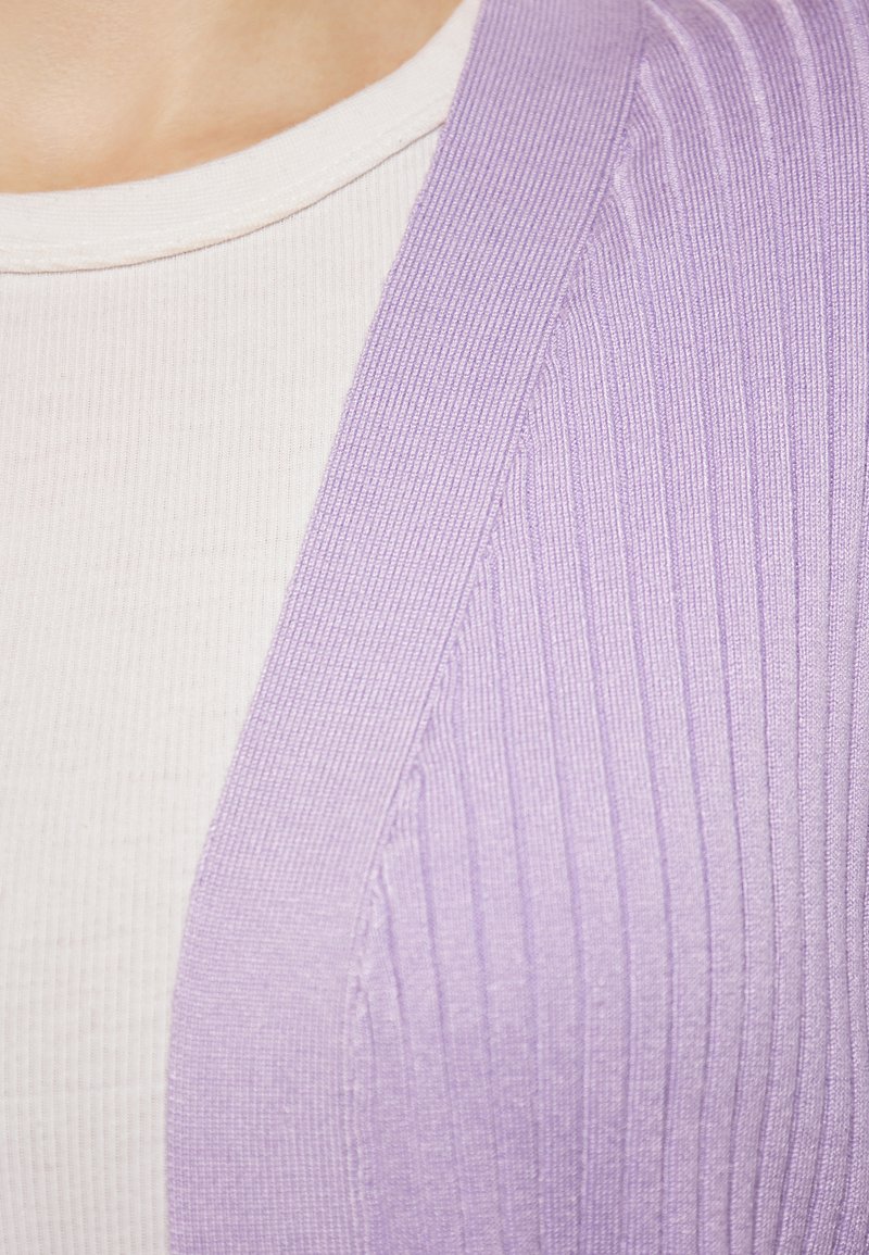 Cardigan rib a lila indossato sopra un top bianco a maniche corte con texture. Il cardigan è realizzato in un morbido tessuto lavorato a maglia con una leggera lucentezza.