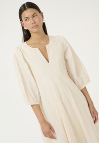 Cremfarbendes Kleid mit V-Ausschnitt, gepufften 3/4-Ärmeln und strukturiertem Stoff. Hat eine fließende Silhouette und horizontale Streifen.