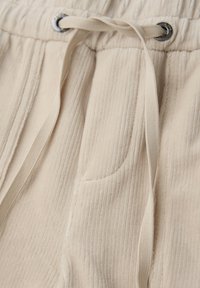 Pantalon en tissu côtelé beige avec taille élastique, poche latérale cousue, œillets métalliques et cordons de serrage beiges.