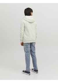 Světle šedá mikina s kapucí na šňůrku, spárovaná se světle modrými džíny a modrými teniskami, viděná zezadu. Celkově hladká textura.