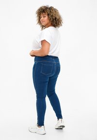 Dark blue denim jeans with a fitted design, elastic waistband, and back pockets. Worn with a white short-sleeve shirt and white sneakers.