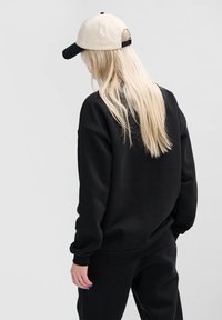 Svart sweatshirt med ribbad mudd och nederkant, bärs med en beige och svart keps. Modellen har långt blont hår. Bakgrunden är enkel grå.