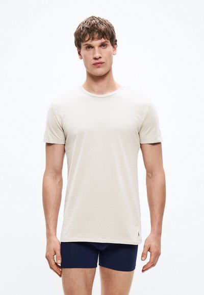 Ανοιχτό μπεζ κοντομάνικο μπλουζάκι με στρογγυλή λαιμόκοψη, κατασκευασμένο από απαλό ύφασμα, με λεπτή υφή και μικρό λογότυπο στο κάτω μέρος.