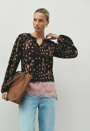 Frau mit blondem Haar, die eine Bluse mit Leopardenmuster und ein rosa Spitzen-Top trägt, hellblaue Jeans, hält eine braune geflochtene Handtasche.