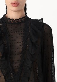 Zwarte doorschijnende blouse met een stippenpatroon, rimpelafwerking, kanten accenten en een hoge kraag. De textuur van de stof creëert diepte en interesse.