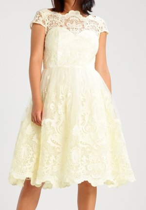 Woman wearing a knee-length cream dress with lace detailing, short sleeves, and a scalloped hem standing against a plain background.