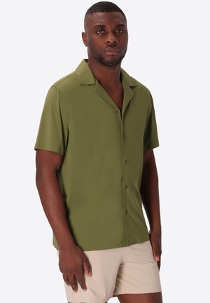 Uomo che indossa una camicia verde oliva a maniche corte con bottoni e pantaloncini beige chiaro, in piedi davanti a uno sfondo bianco semplice.
